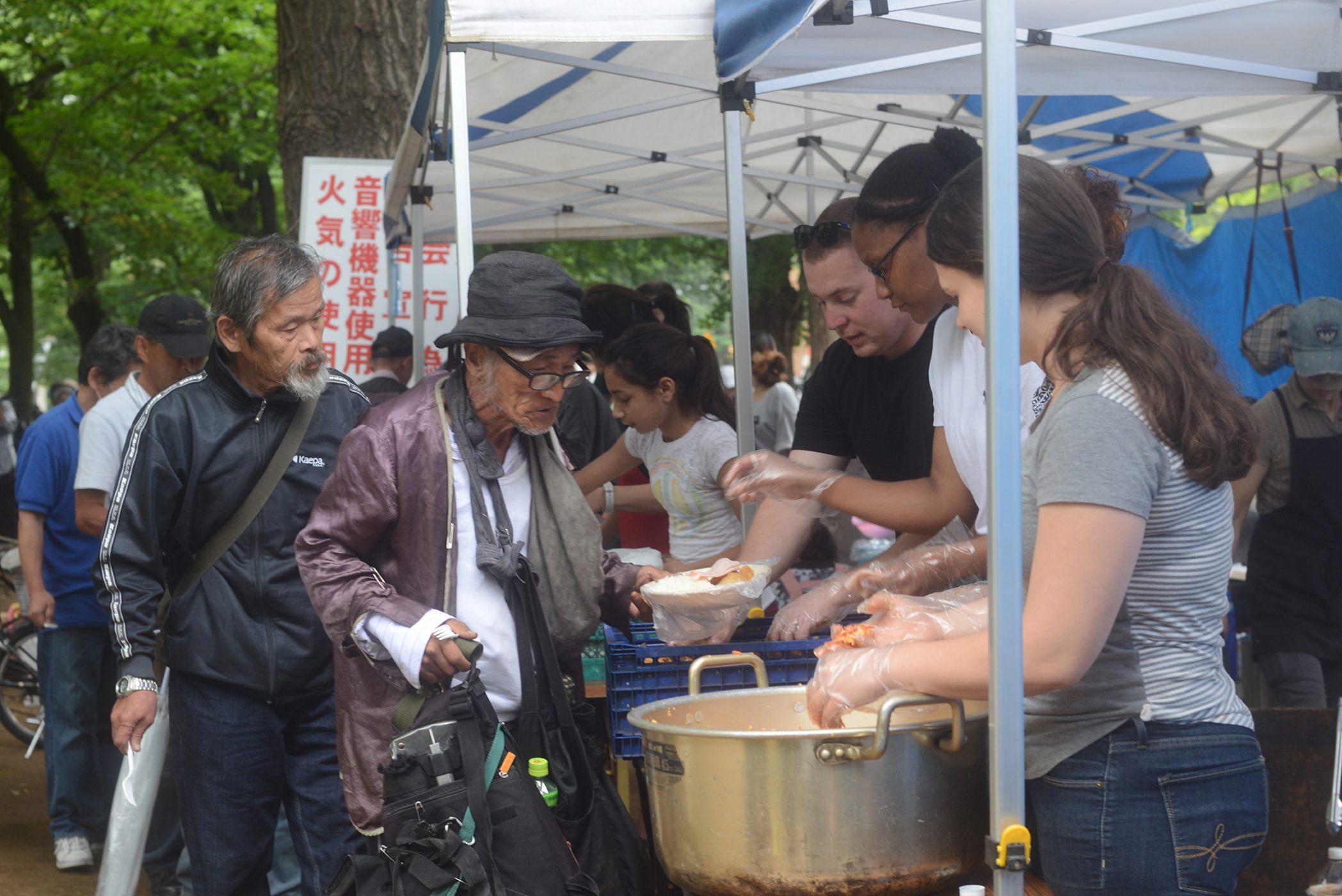 Volunteers serve meals to poor and homeless people in a public park.